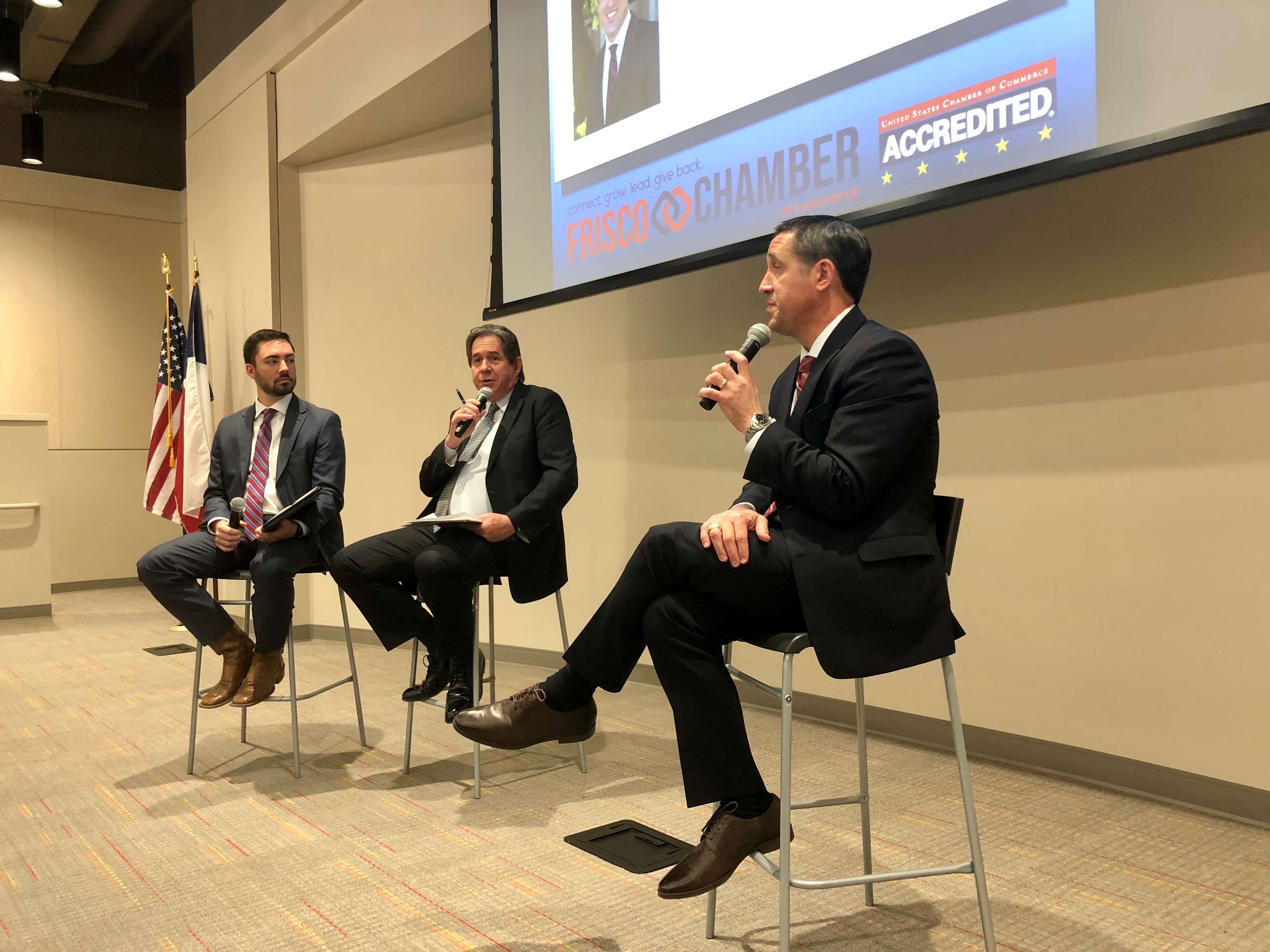 Three representatives from the Frisco Chamber of Commerce sit in chairs at the front of an event. They all hold microphones.