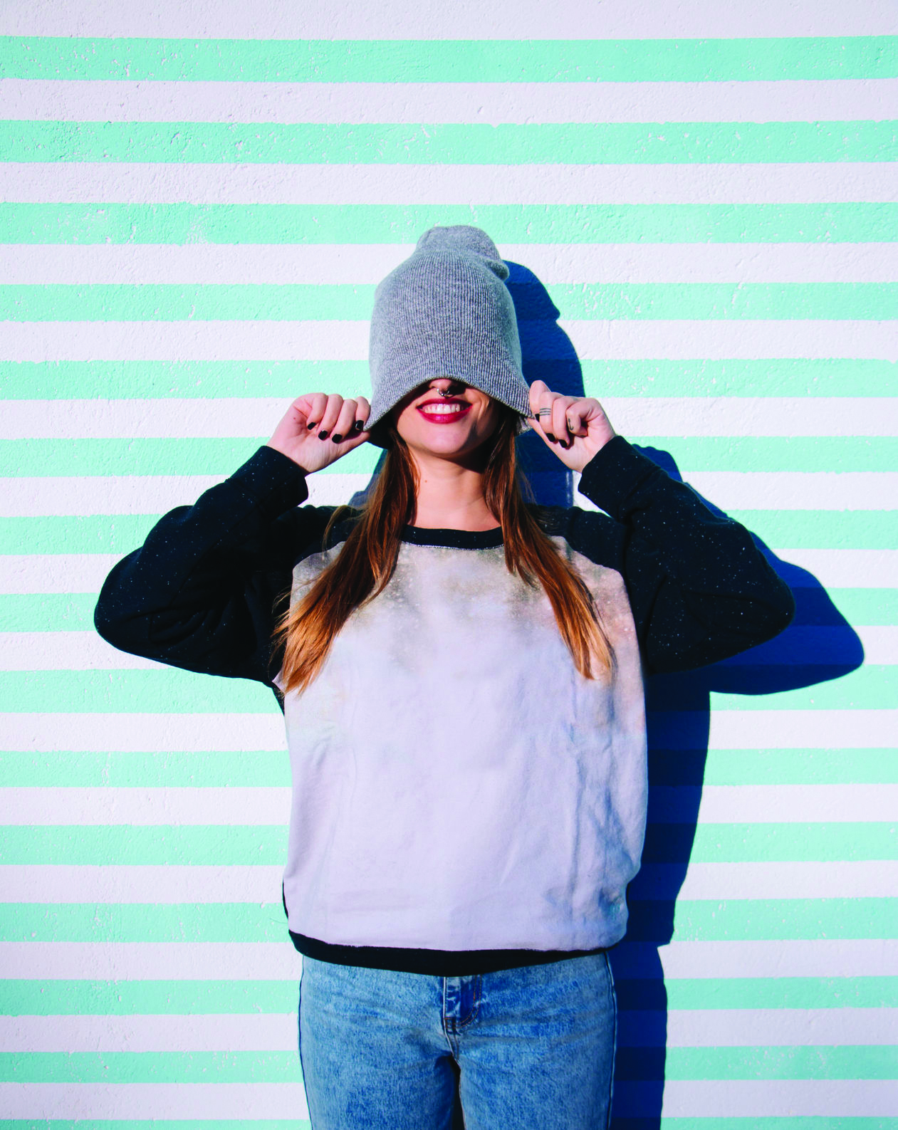 A girl against a striped wall pulls her beanie over her head and smiles.