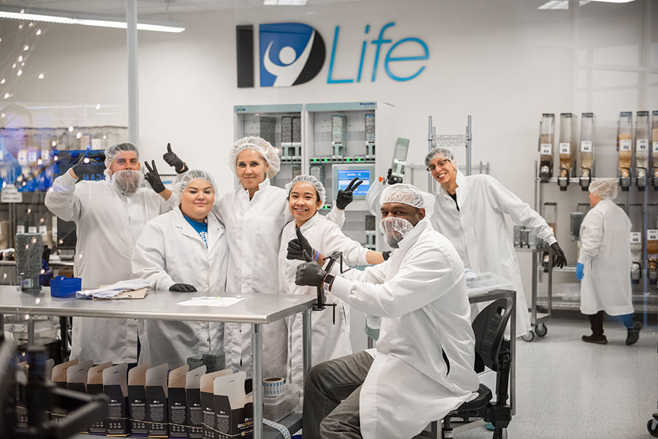 Six lab workers gather for a photo in the lab at ID Life. They wear lab coats and hair nets.